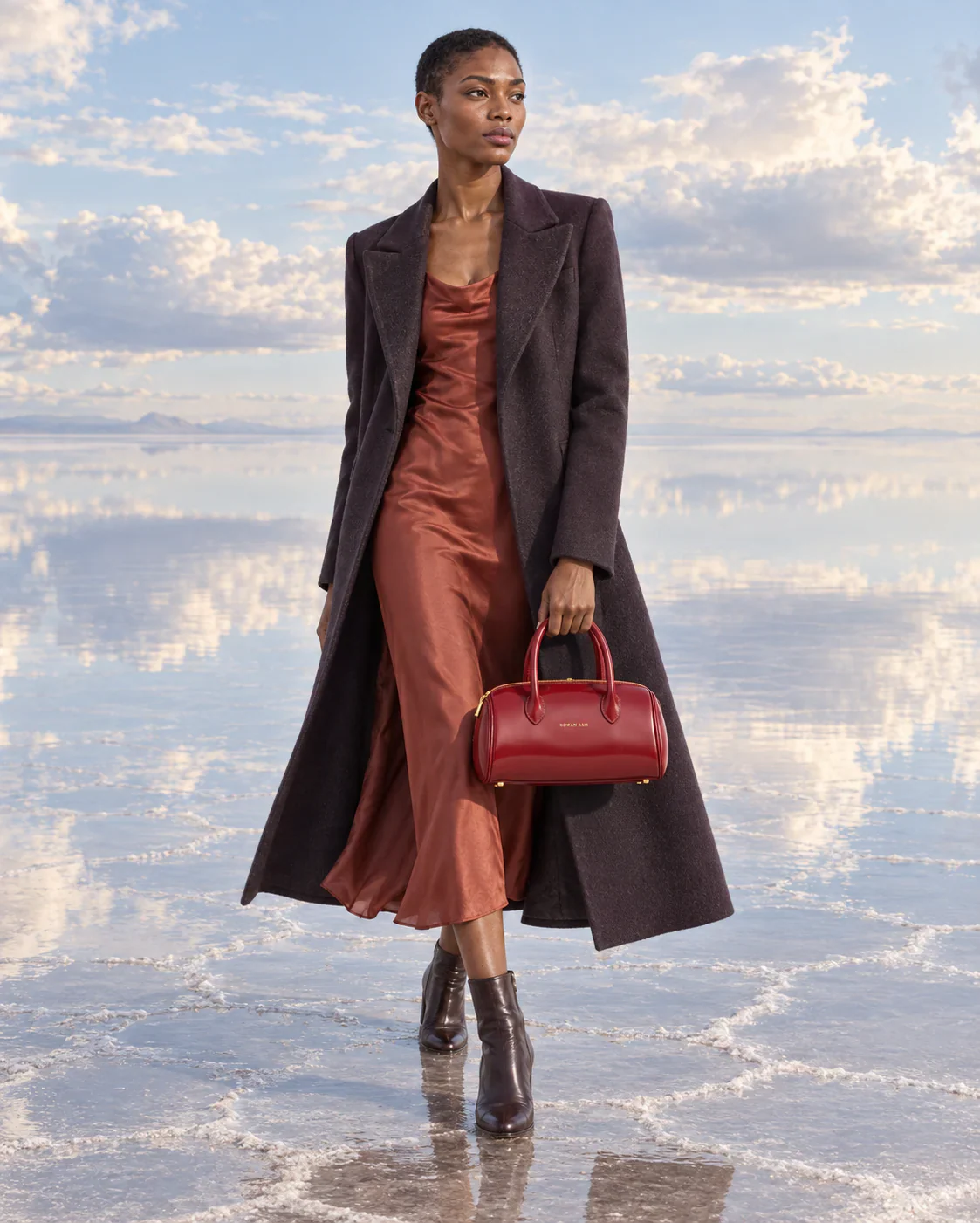 Amara in a brown coat with the bag on a mirrored salt flat